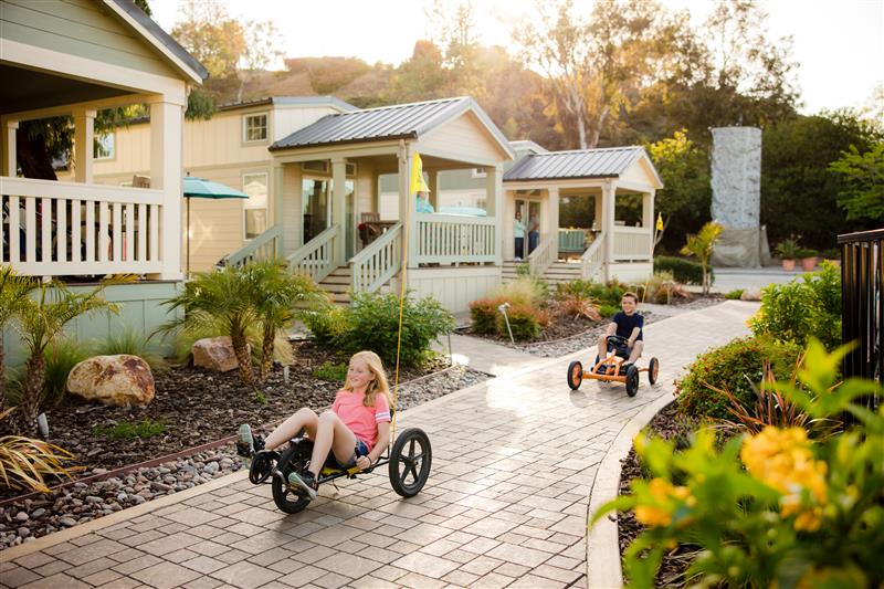 Children ride pedal go-karts along a sunlit brick path past pastel cottages and landscaped gardens.
