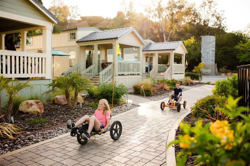 Children ride pedal go-karts on a sunny paved path beside pastel cottages and landscaped gardens.